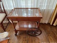 Photo showing full tea cart with glass top and wooden frame including large spoked wooden wheels.