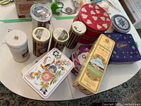 Overview of various food tins and containers including Cadbury, Glenlivet, floral patterned, red heart design, and striped cylinder tins on a white table.