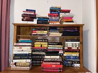 A wide view of multiple stacks of assorted books on a wooden shelf, with various sizes and colors mostly hardcover.