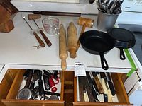 View of counter with two cast iron pans, two wooden rolling pins, meat mallet, glass measuring cup, and utensils in drawer.