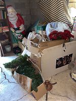 Photo showing large Santa Claus statue behind boxes filled with various Christmas decorations including wreath, faux flowers, and ornaments.