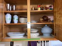 Cabinet shelves with various dishware items including ceramic plates, white bowls, wooden stackable dishes, two ceramic vases with light green gradient, and Revere reproduction pewter bowls along with decorative figurines.