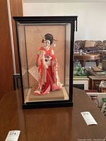 Japanese geisha doll inside a black-framed glass display case with pink floral kimono and fan