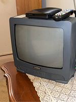 Front angle view of the GoldStar CRT TV set on a wooden table with a lace doily. A black external box and remote control rest on top of the TV.