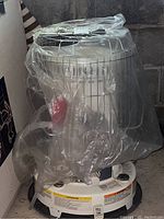 Full view of kerosene heater covered partly in plastic, showing cylindrical shape, metal wire guard, white base, and red control knob.