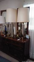 Two silver-tone metal table lamps with off-white drum shades on wooden dresser
