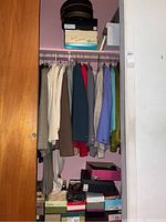 Closet rod with womans pantsuits and jackets in neutral and darker colors, with shoe boxes stored below the hanging clothes and hat boxes above.