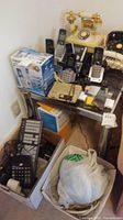 View of collection atop and around a small work desk including rotary phones, cordless phones, calculator, keyboard, and surge protectors