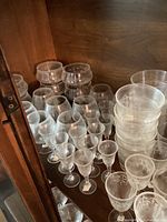 Large collection of assorted stemware, small wine glasses, tumblers, and other glassware inside a wooden cabinet shelf.