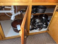 Cabinet with various pots and baking pans stacked inside including stainless steel pots and a large brown cauldron.