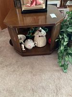 Overall view of an octagonal wooden side table with dark brown finish and woven wicker panels on the sides. Shows open shelf base with some decorative items not included.