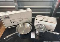 Overview of both cookware items side by side in front of their boxes: skillet on left, saucepan on right.