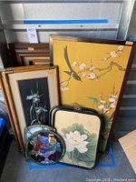 Overall photo showing the assortment of framed artworks including Asian painting with bird, pen and ink floral pieces, lotus flower paintings, family history parchment, and stained glass bowl of flowers and peacock.