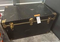 Front and top angled view of the black trunk showing brass corners, studs, and broken hasp/lock area.