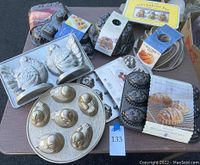 Photo showing assortment of 9 new cake pans including turkey-shaped pans, bundt pans, cakelet molds, gold six-duck cake pan, and silver round pans placed on a table.