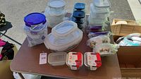 Overview of 23 glass food storage containers of various sizes and shapes with locking plastic lids, arranged on a table outdoors. Some in bubble wrap and original packaging.