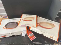 Three boxed Le Creuset pans (black oval baker, orange skillet, small orange oval baker) displayed on surface