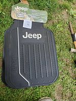 Photo of new black Jeep rubber floor mat with Jeep logo and packaging visible in outdoor setting