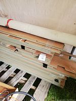 Stack of light brown laminate flooring planks with wood grain pattern and tongue and groove edges, stacked on a pallet with a white linoleum roll placed on top.