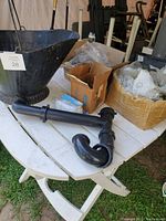 Large black plastic ash bucket, black plastic drain pipe on white plastic table with cardboard boxes holding various small parts