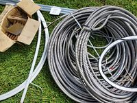 Photo showing coil of flexible metal conduit with white plastic tubing and a cardboard box of metal fasteners on grass.