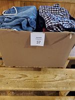 Cardboard box with folded clothing items including denim jeans and checkered shirts visible