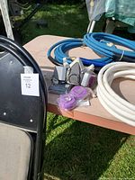 Two blue high pressure hoses, white trailer hose, gray vent mask with purple filters, and parts of black folding chairs on a wooden table outdoors