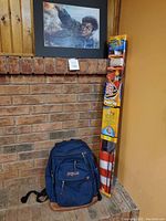 Front view of blue JanSport backpack placed against brick wall with Harry Potter kite standing next to it, under a Harry Potter character painting.
