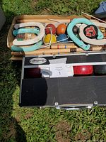 View of box containing rubber horseshoes, croquet mallets and balls, and a black bocce ball case on grass.