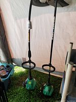 Two gas-powered green weed eaters with black handles and circular guards, positioned upright on grass with a tent or tarp background.