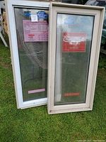 Two fixed rectangular windows with white vinyl frames resting on grass, brand stickers visible on glass.