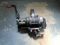 Full view of the Warn 12 volt winch showing motor, mounting base, steel cable winding spool, and electrical cords with red connector.