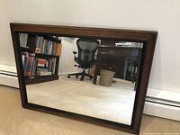 Front view of rectangular wall mirror with brown wood frame and black inset, placed on carpet showing reflection of room furniture.