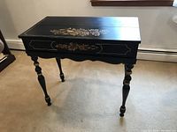 Full view of black antique flip top writing desk featuring hand-painted floral and fruit decoration on front and top, decorative gold borders, turned legs with gold accents.