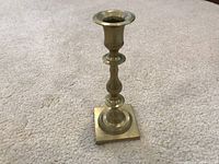 Front view of single vintage brass tapered candlestick standing upright on carpeted floor.