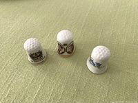 Photo of three bone china thimbles showing the side images and white dimpled tops on green fabric surface.