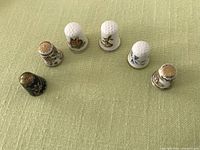Top view of six decorative thimbles displayed on green fabric background, showing enamel and bone china types with floral and figure designs.