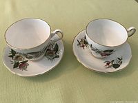 Two fine china tea cup and saucer sets displayed on a green surface, showing flower designs and gold trim.