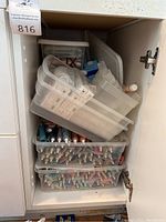 Photo of white cabinet door open revealing stacked plastic bins containing assorted paint tubes and art supplies including crayons and pencils.