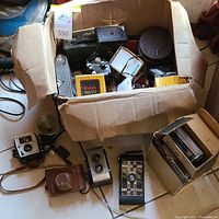 Box filled with assorted vintage cameras including Kodak Kodaflex and others, camera cases, camera accessories, and a camera flash unit.