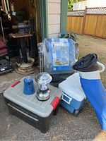 Multiple camping items including cooler, camping kettle, camping lantern, storage container, and camping chair in bag grouped outside.
