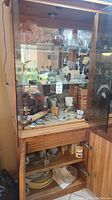 Overall view of wood and glass cabinet holding assorted glassware, mugs, platters, bowls, and collectible items arranged on shelves