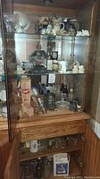 Wide view showing three shelves of collectibles in a wooden cabinet including glassware, bowls, shot glasses, steins, figurines, and other small items.