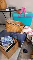 View of Longaberger basket, turquoise plastic tote with pink handles, blue tote with t-shirts, Dodge Viper model kit box, and Coca-Cola cushion on floor near table