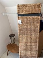 View of rattan room divider panels stacked and leaning against a wall in a corner, showing woven natural material and metal frame straps, with small table and mosaic stand visible nearby.