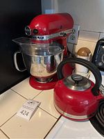 Red KitchenAid Artisan stand mixer with stainless steel bowl and splash guard next to a red kettle on kitchen countertop
