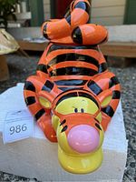 Front view of ceramic Tigger cookie jar depicting Tigger lying on his back, glossy orange with black stripes, face with pink nose and yellow cheeks.