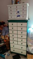Smaller white chest with red knobs on top of larger green and white chest with green knobs, placed near other craft and storage items.