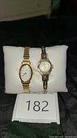Pair of watches displayed on white cushion, Timex on left gold-tone, floral watch on right with black embroidered band.