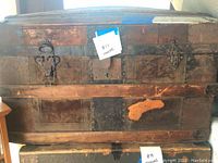 Front view of antique wooden trunk showing metal locking clasps, handles, and areas of damage.